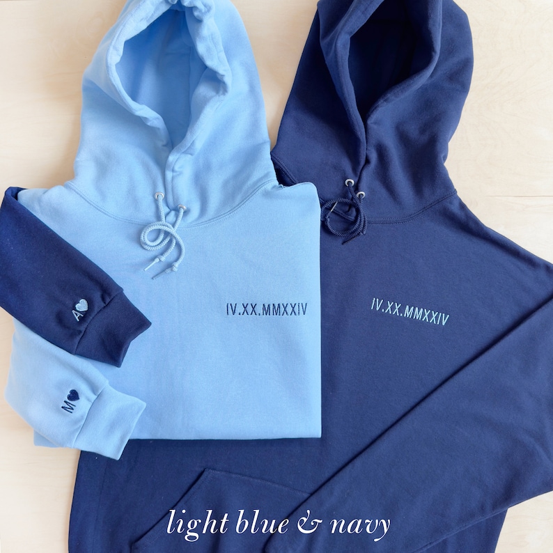 K&ouml;nnte beinhalten: Zwei Kapuzenpullover, einer hellblau und einer marineblau. Der hellblaue Pullover hat einen marineblauen &Auml;rmel mit Initialen und einem Herz. Beide Pullover haben das r&ouml;mische Datum "IV.XX.MMXXIV" auf der Vorderseite. Der Text "light blue & navy" steht unten.