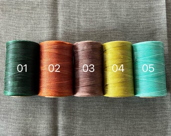 Puede incluir: Cinco carretes de hilo de diferentes colores. Los carretes están etiquetados con los números 01, 02, 03, 04 y 05. Los colores son verde oscuro, naranja, marrón, amarillo y turquesa.