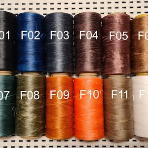 Puede incluir: Una colección de doce carretes de hilo en varios colores, incluyendo negro, azul marino, marrón, naranja y blanco. Cada carrete está etiquetado con un número "F" correspondiente, que va de F01 a F12.