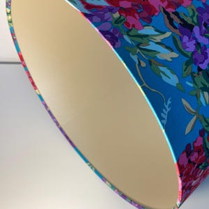 Op de afbeelding: Een close-up van een lampenkap met een bloemenpatroon. De lampenkap is gemaakt van stof en heeft een cr&egrave;mekleurige voering. Het bloemenpatroon is in tinten roze, paars en blauw op een turquoise achtergrond.