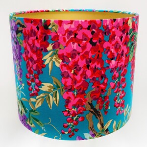 Op de afbeelding: Een turquoise blauwe lampenkap met een bloemenpatroon van roze en paarse wisteria bloemen. De lampenkap heeft een gouden binnenvoering.