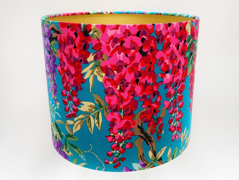 Op de afbeelding: Een turquoise blauwe lampenkap met een bloemenpatroon van roze en paarse wisteria bloemen. De lampenkap heeft een gouden binnenvoering.