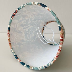 May include: A white lampshade with a floral pattern in shades of blue, green, and orange. The lampshade is made of fabric and has a white lining.