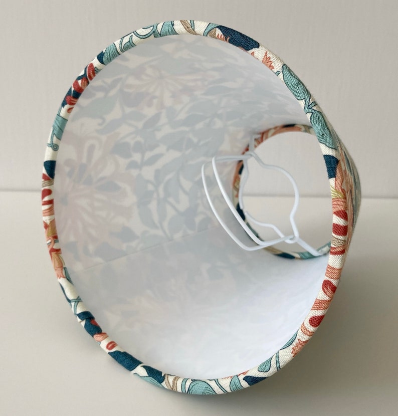 May include: A white lampshade with a floral pattern in shades of blue, green, and orange. The lampshade is made of fabric and has a white lining.