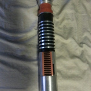 Puede incluir: Una empuñadura de sable de luz plateada con un agarre negro y plateado y un emisor rojo y plateado. La empuñadura tiene una serie de anillos plateados y una sección roja y plateada en el medio.