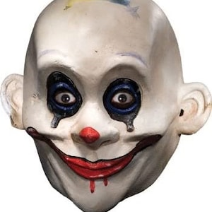Puede incluir: Una máscara de payaso blanca con maquillaje azul para los ojos, una sonrisa roja y una nariz roja. La máscara tiene un aspecto desgastado, con salpicaduras de pintura y grietas.