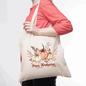 May include: A white canvas tote bag with a design of three pumpkins, leaves, and berries. The text "Happy Thanksgiving" is printed in brown on the bag.