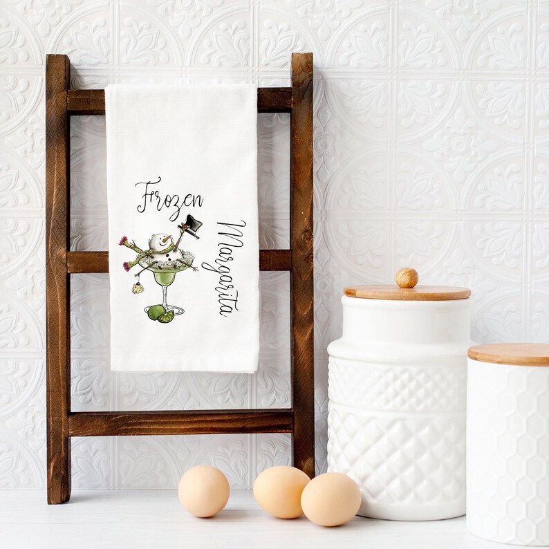 May include: A white kitchen towel featuring a graphic of a snowman sitting in a margarita glass with the text Frozen Margarita. The towel is draped over a dark brown wooden ladder rack, with three light brown eggs and white canisters nearby.