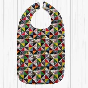 May include: A colorful bib with a patchwork design. The bib features a variety of fabrics in red, green, yellow, and black, arranged in a geometric pattern. It has a snap closure at the neck and is designed to catch spills.