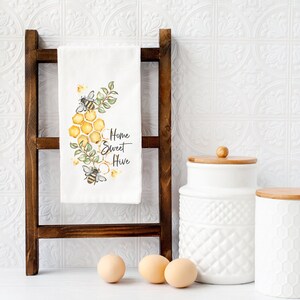 May include: A white kitchen towel with a bee and honeycomb design, hanging on a dark brown wooden ladder. The towel has the words "Home Sweet Hive" printed on it. Three eggs and two white ceramic jars with wooden lids are in the foreground.