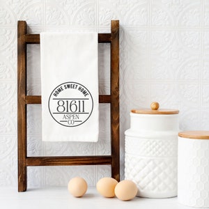May include: White kitchen towel with a black circle design that says "Home Sweet Home 81611 Aspen CO" hanging on a wooden ladder.