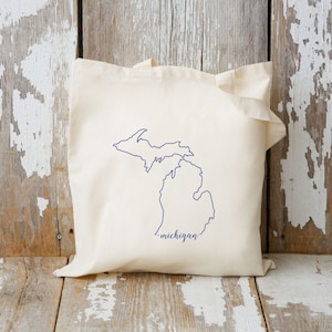 Peut inclure: Un sac fourre-tout crème avec un contour bleu marine de l'État du Michigan. Le mot "michigan" est écrit en cursive en dessous du contour de l'État. Le sac est fait d'un tissu doux.