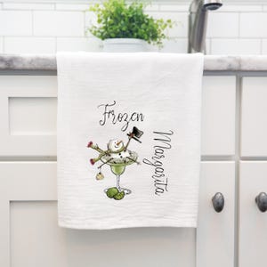 May include: A white kitchen towel featuring a graphic of a snowman wearing a black top hat and a green scarf, sitting inside a green margarita glass. The text "Frozen Margarita" is printed in black script lettering next to the illustration.
