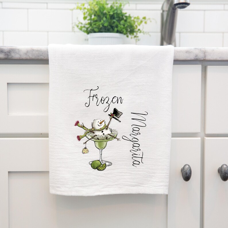 May include: A white kitchen towel featuring a graphic of a snowman wearing a black top hat and a green scarf, sitting inside a green margarita glass. The text "Frozen Margarita" is printed in black script lettering next to the illustration.