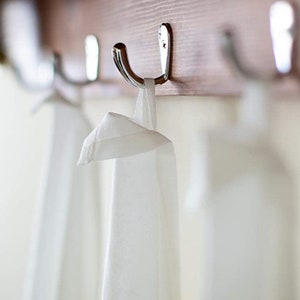 May include: A plain white fabric towel with a hanging loop suspended from a polished silver metal hook. The hook is securely attached to a horizontal wooden board. The towel hangs vertically, displaying a smooth, clean, and simple textile surface.