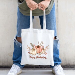 May include: White canvas tote bag with a watercolor design of pumpkins, leaves, and berries. The text "Happy Thanksgiving" is printed in brown below the design.