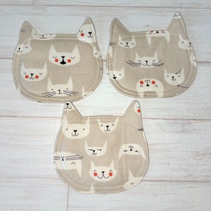 May include: Three beige fabric cat-shaped coasters with black and white cat faces and red accents. The coasters are decorated with a variety of cat faces, some with closed eyes and some with open eyes.