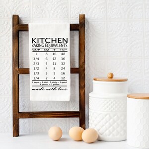 May include: White kitchen towel with black text that reads "Kitchen Baking Equivalents" and includes a chart of cup, ounce, tablespoon, and teaspoon conversions. The text at the bottom reads "2 cups = 1 pint, 2 pints = 1 quart, 4 quarts = 1 gallon, made with love".
