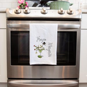 May include: A white kitchen towel featuring a graphic of a snowman sitting in a margarita glass with the text Frozen Margarita. The snowman wears a black top hat and green scarf, with two lime wedges at the base of the glass stem on the fabric.