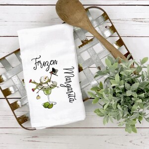May include: A white kitchen towel featuring a graphic of a snowman inside a margarita glass with the text Frozen Margarita. The snowman wears a black top hat and a green scarf, with lime wedges at the base of the glass and a small ornament hanging.
