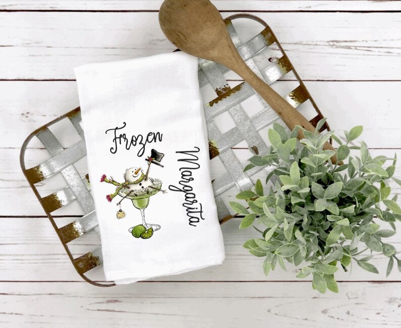 May include: A white kitchen towel featuring a graphic of a snowman inside a margarita glass with the text Frozen Margarita. The snowman wears a black top hat and a green scarf, with lime wedges at the base of the glass and a small ornament hanging.