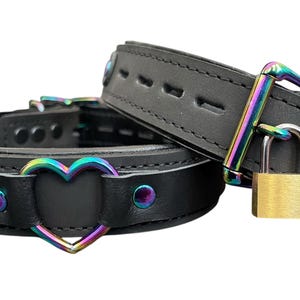 May include: Two black leather collars with rainbow-colored metal hardware. One collar has a heart-shaped cutout and the other has a padlock.