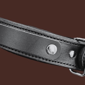 May include: Close-up of a black leather strap with silver-colored metal rivets and a buckle. The strap is stitched with white thread along the edges. The background is a solid brown color.