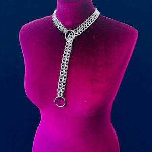 Op de afbeelding: Een zilverkleurige ketting met een dubbelstrengs ontwerp. De ketting heeft een grote ring in het midden en een kleinere ring aan de onderkant. Het wordt getoond op een magenta mannequin tegen een donkerblauwe achtergrond.