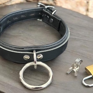May include: Black leather collar with a silver metal ring and buckle. Includes a small gold padlock and two keys. The collar has a white accent stripe and is secured with rivets.