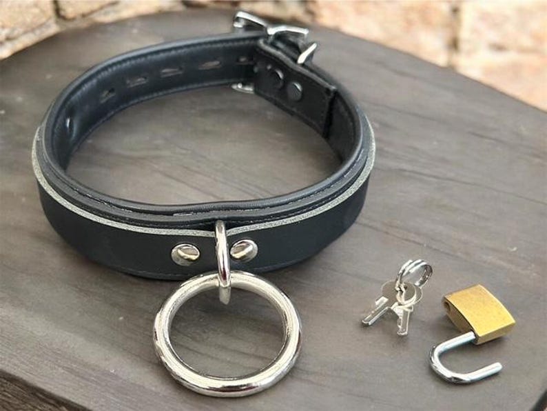 May include: Black leather collar with a silver metal ring and buckle. Includes a small gold padlock and two keys. The collar has a white accent stripe and is secured with rivets.