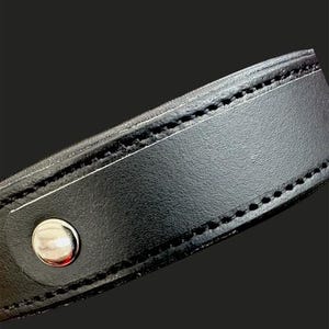 May include: Close-up of a black leather collar with a silver-colored snap closure. The collar is stitched with black thread along the edges. The background is a solid black.