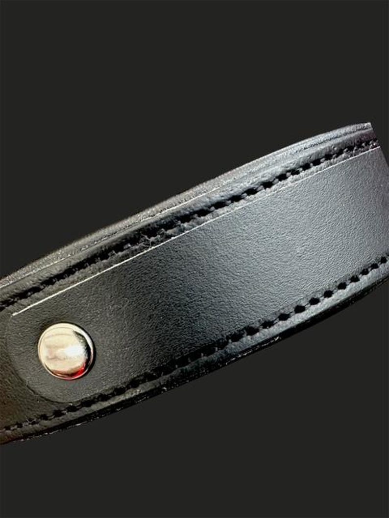May include: Close-up of a black leather collar with a silver-colored snap closure. The collar is stitched with black thread along the edges. The background is a solid black.