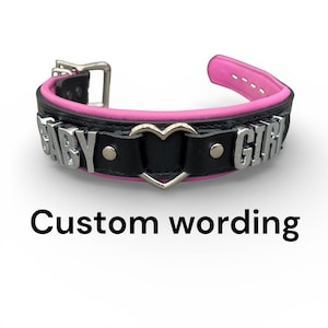 May include: Black and pink leather collar with silver-toned metal hardware. The collar features the words "BABY GIRL" in silver letters and a heart-shaped accent. The text "Custom wording" is below the collar.