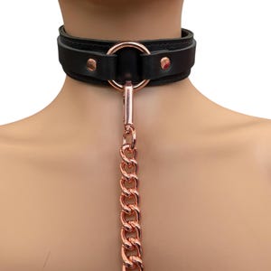 May include: A black leather choker with a rose gold metal ring and chain. The chain is attached to the ring and hangs down from the choker.
