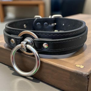 Op de afbeelding: Zwarte leren halsband met een grote zilveren metalen ring en gesp. De halsband is gewatteerd en heeft een gestikt ontwerp. De metalen ring is met een kleinere metalen ring aan de halsband bevestigd. De halsband ligt op een houten oppervlak.