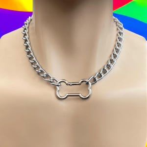 May include: A silver-colored chain necklace with a bone-shaped pendant. The necklace is made of thick, interlocking links and is displayed on a mannequin. The pendant is centered and has a smooth, polished finish.