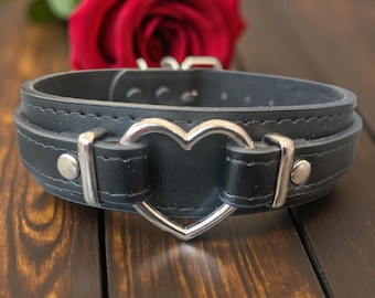 May include: Black leather choker with a silver heart-shaped center. The choker has a double-layered design with silver hardware and a buckle closure. A red rose is in the background.