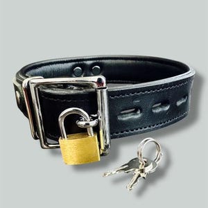 May include: Black leather collar with a silver buckle and a gold padlock. Two silver keys are next to the collar. The collar is thick and has stitching along the edges.