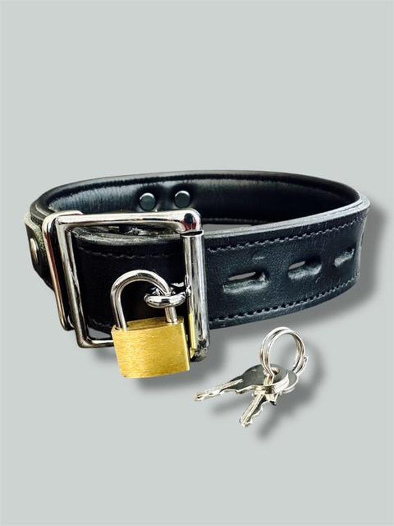 May include: Black leather collar with a silver buckle and a gold padlock. Two silver keys are next to the collar. The collar is thick and has stitching along the edges.