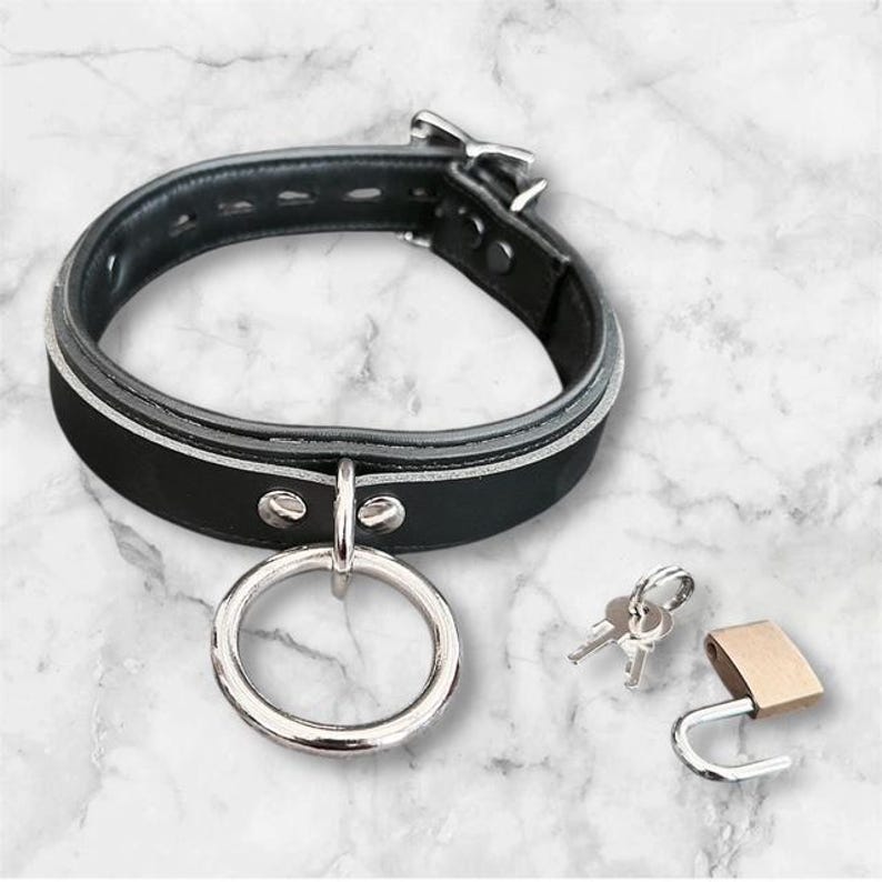 May include: Black leather collar with a silver ring and buckle. Includes a small gold padlock and two keys. The collar has a white stripe and is designed with a classic aesthetic.