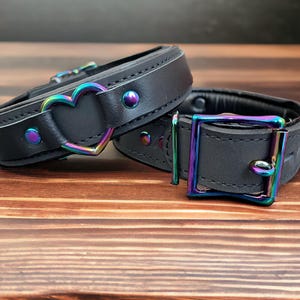 May include: Two black leather collars with iridescent metal hardware. One collar features a heart-shaped buckle, while the other has a rectangular buckle.