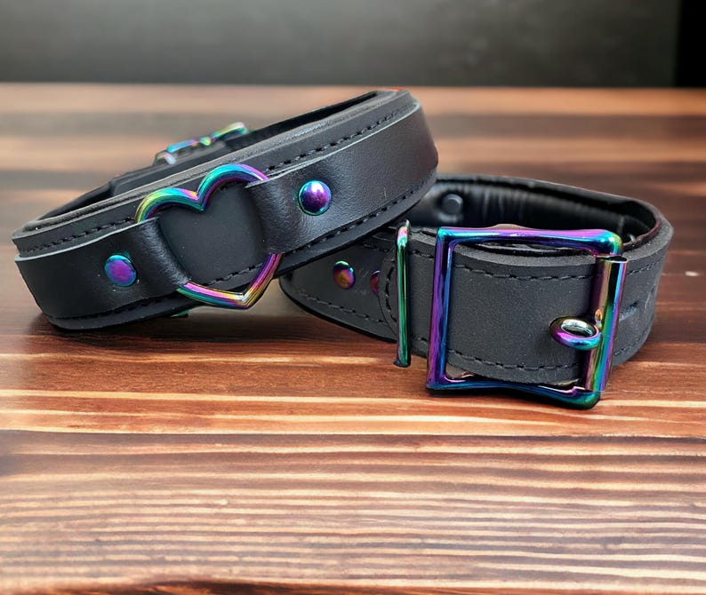 May include: Two black leather collars with iridescent metal hardware. One collar features a heart-shaped buckle, while the other has a rectangular buckle.