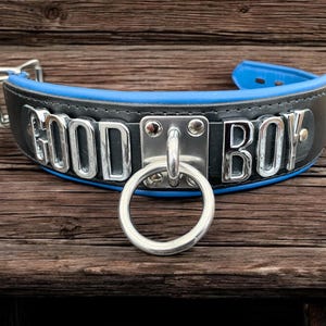 May include: A black leather collar with a blue trim and silver metal accents. The collar has the words "GOOD BOY" embossed in silver metal letters.