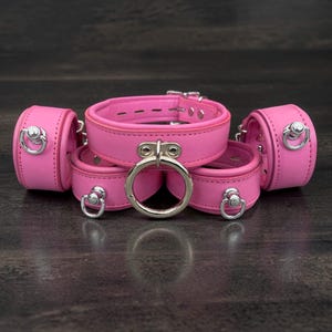 Op de afbeelding: Een set roze leren boeien, waaronder een halsband, polsboeien en enkelboeien. Elk stuk heeft zilverkleurige metalen hardware, inclusief ringen en gespen. De items zijn gerangschikt op een donker, getextureerd oppervlak.