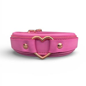 May include: A pink leather collar with a heart-shaped gold-colored metal accent. The collar has a buckle closure and two small gold-colored studs. The design is simple and elegant.