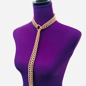 Op de afbeelding: Goudkleurige ketting met een dubbelstrengs ontwerp rond de nek, verbonden met een enkele, lange ketting die naar beneden hangt. De ketting wordt getoond op een paarse mannequin.