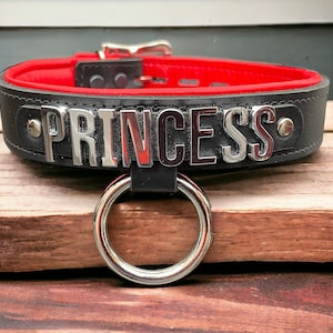 May include: Black leather collar with red lining and a silver metal ring. The collar has the word "PRINCESS" spelled out in silver metal letters.