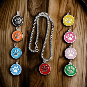 May include: A silver chain necklace with a red paw print pendant. The image also shows several other colourful paw print pendants in black, orange, blue, yellow, pink, and green. The pendants are round with a silver border.