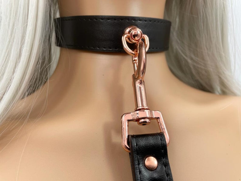 May include: A black leather choker with a rose gold metal clasp and ring. The choker is worn on a person's neck.
