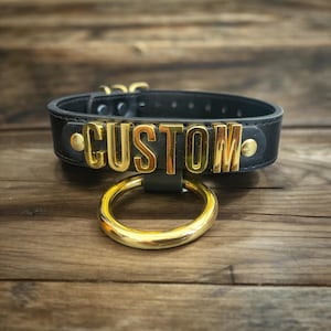 Puede incluir: Gargantilla de cuero negro con un anillo de metal dorado y la palabra "CUSTOM" escrita en letras doradas.